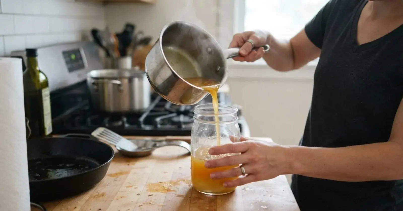 Person pouring hot liquid from a saucepan into a glass jar in a kitchen