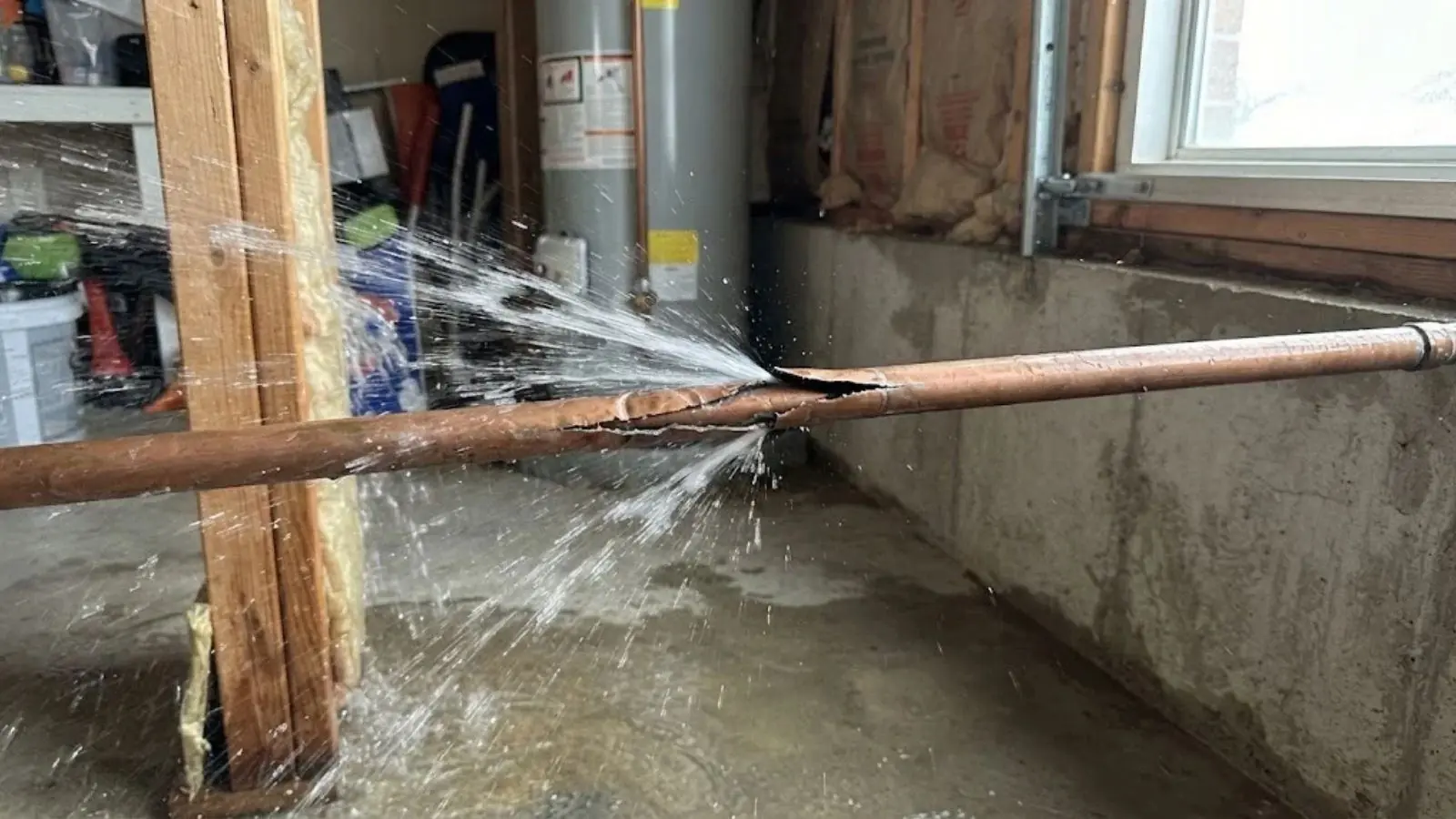 Split copper pipe spraying water across a basement beside a water heater